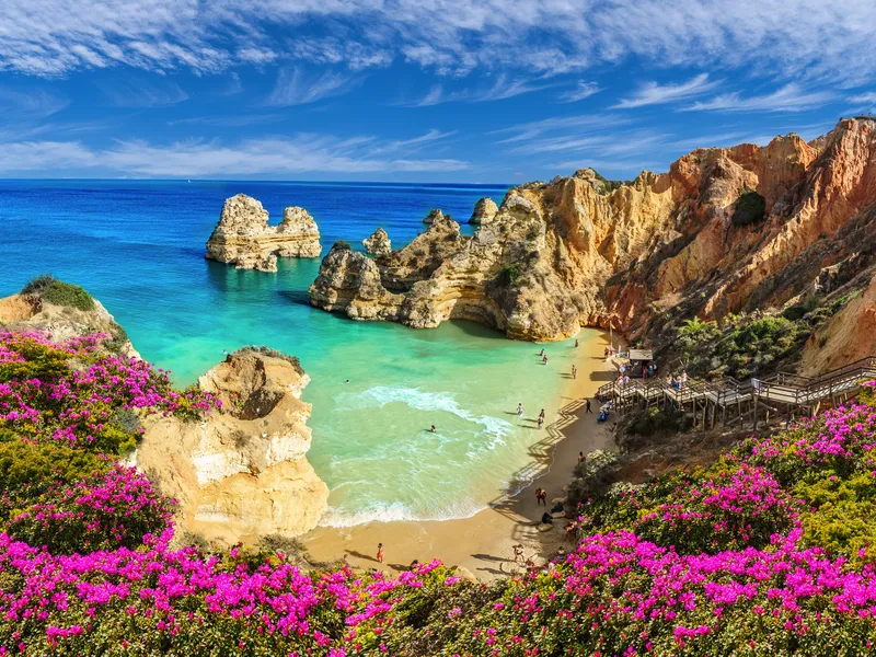 Küstenlandschaft mit Felsen, türkisfarbenem Meer und rosa Blumen im Vordergrund.