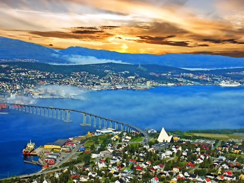 Stadtansicht mit Brücke, Häusern und Bergen bei Sonnenuntergang.