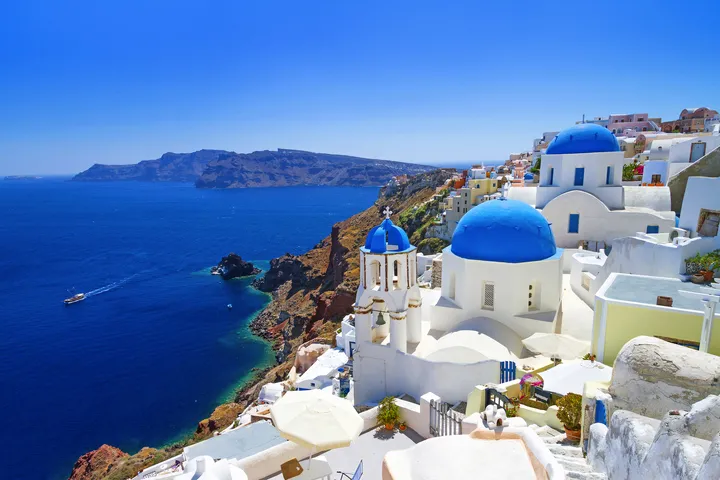 Weiße Gebäude mit blauen Kuppeln auf Santorini, Meer im Hintergrund.