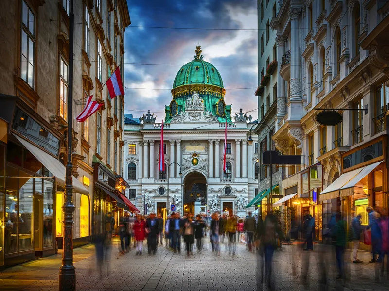 Menschen auf belebter Straße vor barockem Gebäude in Wien bei Dämmerung.