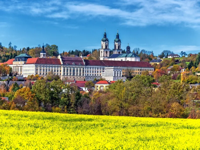 Barocke Klosteranlage mit Türmen, umgeben von Herbstbäumen und gelbem Blumenfeld.