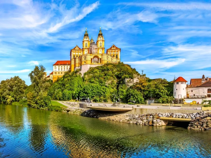 Barockes Kloster auf Hügel über Fluss, umgeben von Bäumen und blauem Himmel.