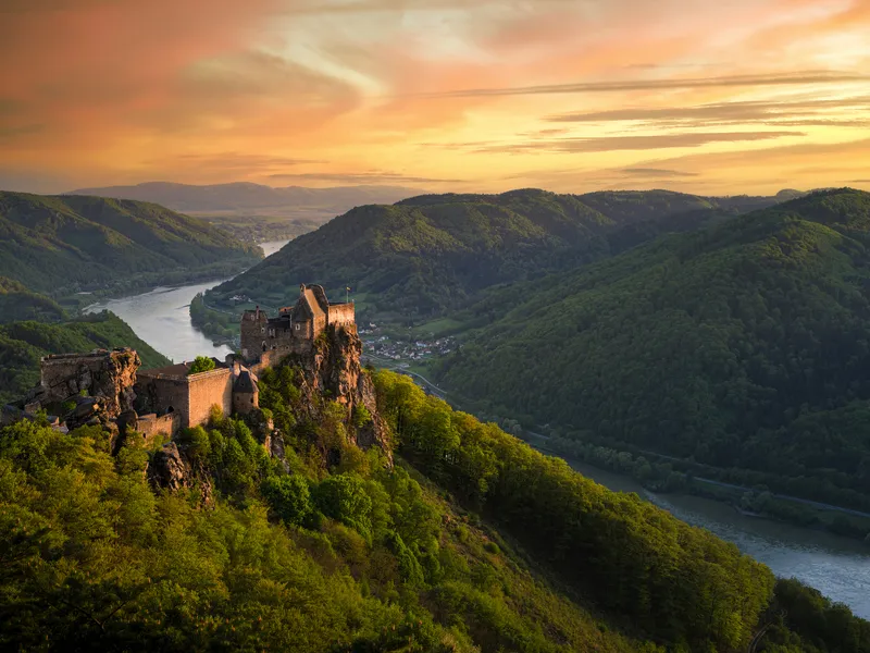Burgruine auf Hügel mit Fluss in malerischer Hügellandschaft bei Sonnenuntergang.
