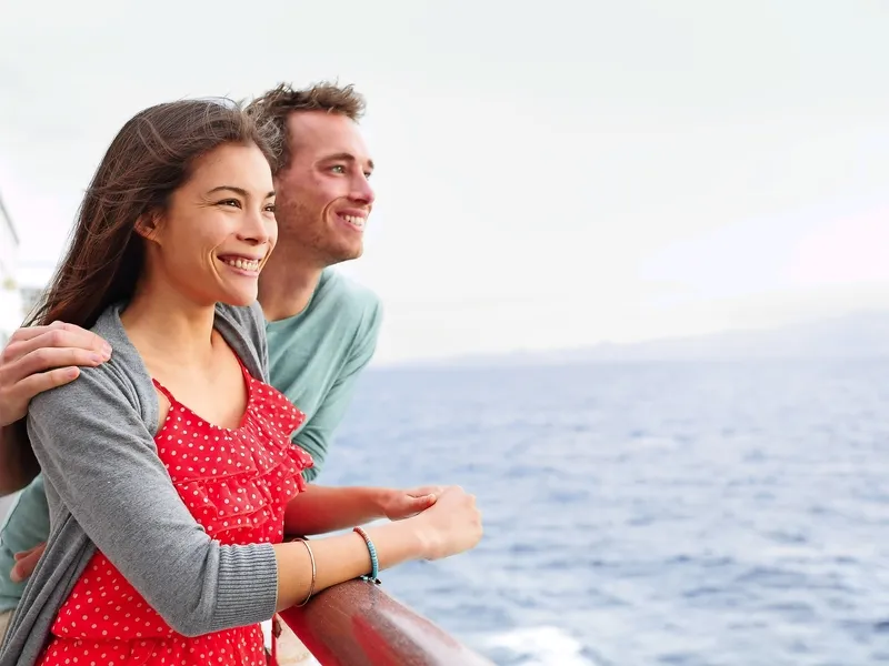 Ein junges Paar steht auf dem Deck eines Kreuzfahrtschiffs und blickt lächelnd aufs Meer.
