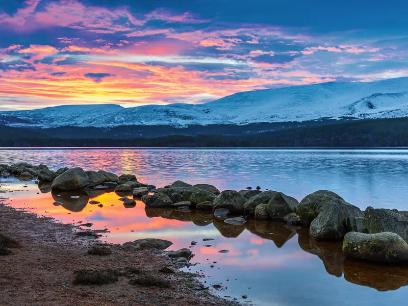 Sonnenaufgang über Loch Morlich im Cairngorms-Nationalpark, Schottland, mit schneebedeckten Bergen im Hintergrund und einem ruhigen See im Vordergrund.