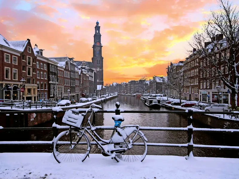 Fahrrad mit Schnee bedeckt steht auf einer Brücke mit Blick auf einen verschneiten Kanal in einer europäischen Stadt bei Sonnenuntergang.