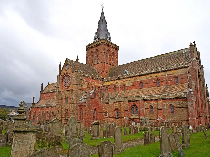 Eine historische, rote Backsteinkathedrale mit mehreren Dächern und einem hohen Turm, umgeben von alten Grabsteinen auf einem Friedhof unter einem bewölkten Himmel.