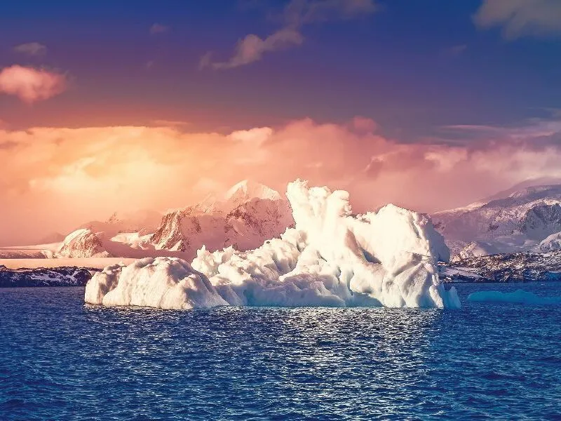 Eine beeindruckende Landschaft mit einem großen, schneebedeckten Eisberg, der im blauen Wasser treibt, im Hintergrund eine Gebirgskette und ein farbenfroher Himmel im Sonnenuntergang.