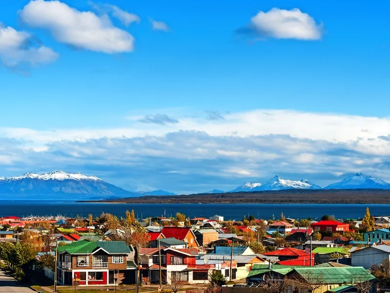 Bunte Häuser in einer kleinen Stadt mit einem See im Hintergrund und schneebedeckten Bergen unter einem blauen Himmel mit weißen Wolken.