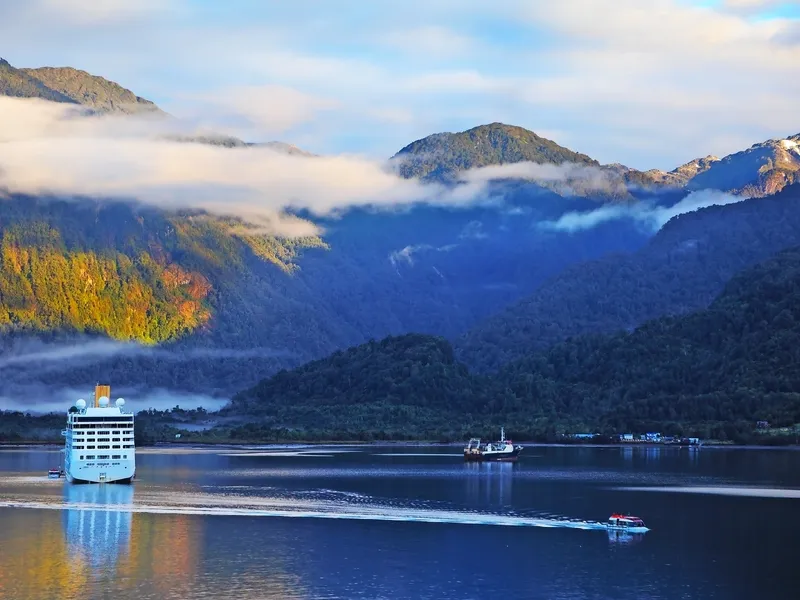 Ein Kreuzfahrtschiff fährt durch eine ruhige Bucht, umgeben von bewaldeten Bergen, die teilweise von Nebel und Wolken bedeckt sind. Im Vordergrund ist ein kleineres Boot auf dem Wasser zu sehen.