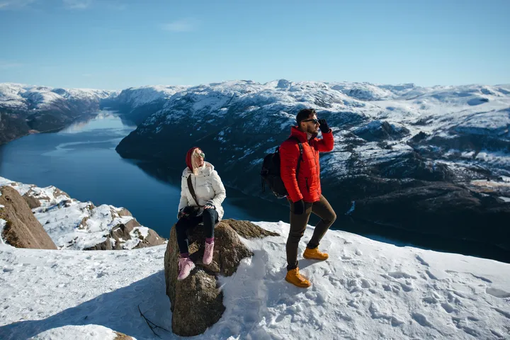 Ein Mann und eine Frau genießen die Aussicht auf schneebedeckte Berge und einen Fjord. Die Frau sitzt auf einem Felsen, während der Mann steht. Beide tragen Winterkleidung.