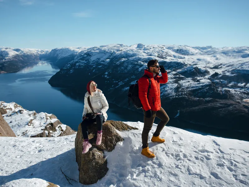 Ein Mann und eine Frau genießen die Aussicht auf schneebedeckte Berge und einen Fjord. Die Frau sitzt auf einem Felsen, während der Mann steht. Beide tragen Winterkleidung.