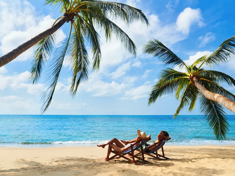 Zwei Personen entspannen auf Liegestühlen am Strand unter Palmen, mit Blick auf das blaue Meer und den klaren Himmel.
