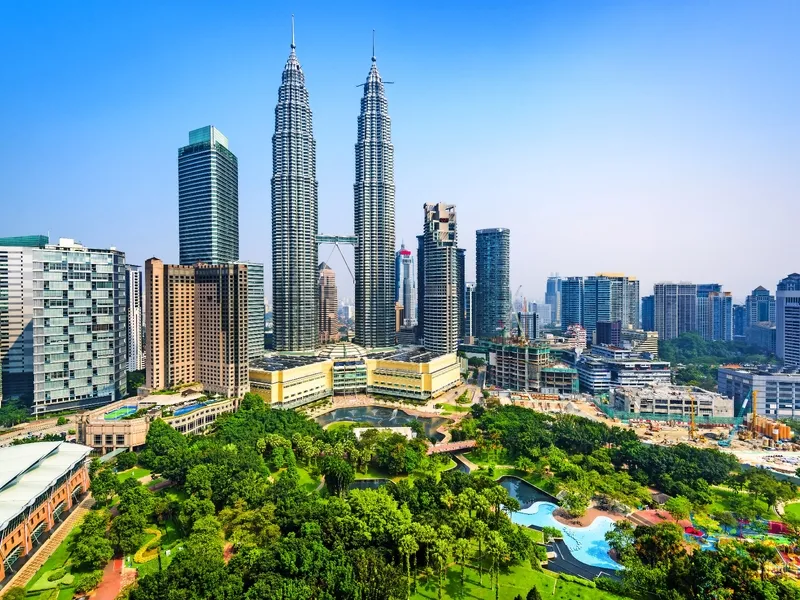 Skyline von Kuala Lumpur mit den Petronas Towers und umliegenden modernen Gebäuden sowie einem grünen Park im Vordergrund bei klarem Himmel.