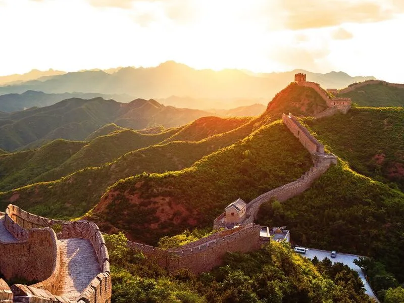 Chinesische Mauer bei Sonnenuntergang, umgeben von grünen Hügeln und Bergen.
