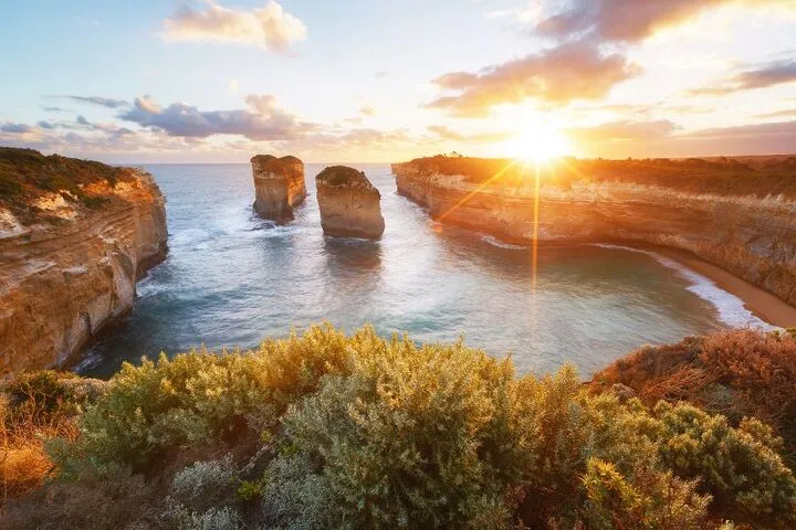 Sonnenuntergang über der Küste mit Felsen im Meer und bewachsenen Klippen im Vordergrund.