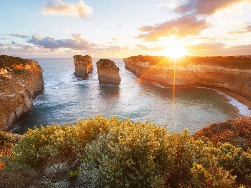 Sonnenuntergang über der Küste mit Felsen im Meer und bewachsenen Klippen im Vordergrund.