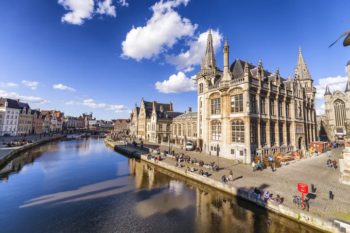 Blick auf die Stadt Gent in Belgien mit der Sint-Niklaaskerk und dem Fluss Leie bei sonnigem Wetter.