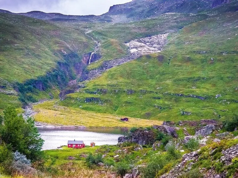 Kleines rotes Haus vor einer grünen, bergigen Landschaft mit Wasserfall.