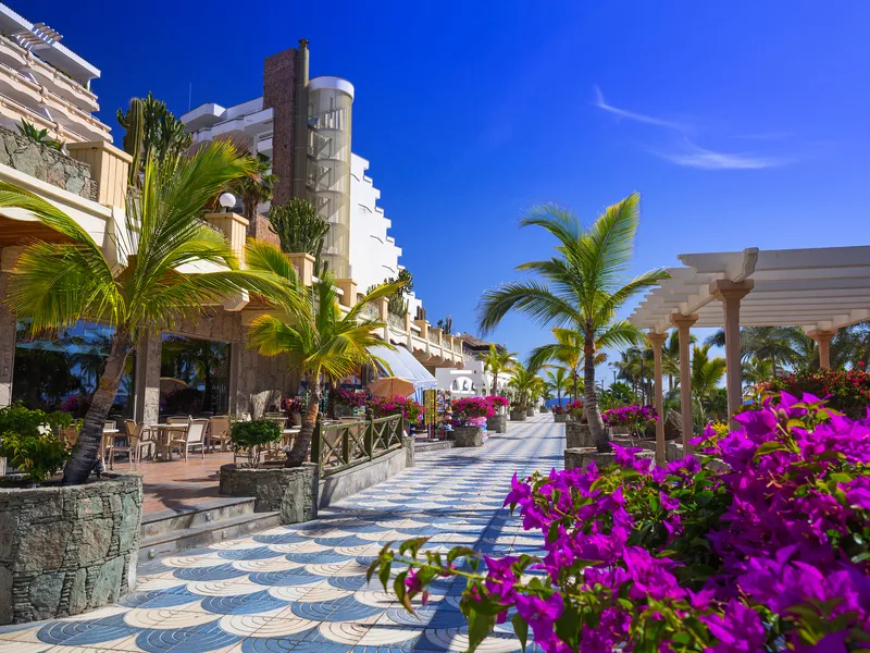 Promenade in Taurito auf Gran Canaria mit Palmen, Blumen und Gebäuden bei sonnigem Wetter.