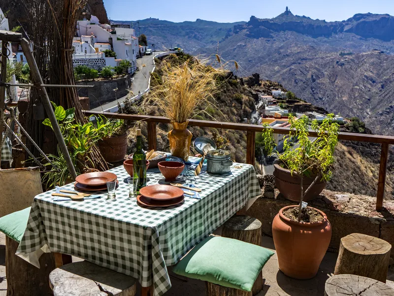Tisch mit kariertem Tischtuch, Geschirr und Pflanzen auf Terrasse mit Bergblick.
