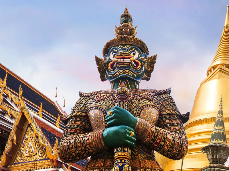 Thailändischer Tempel mit goldener Pagode und großer Wächterstatue.