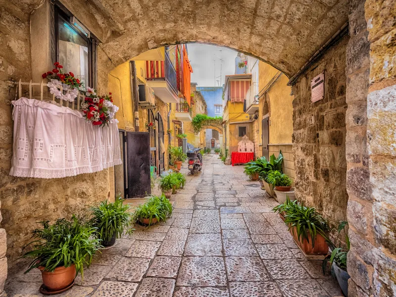 Steinbogen über Gasse mit Pflanzen und bunten Gebäuden in mediterranem Dorf.