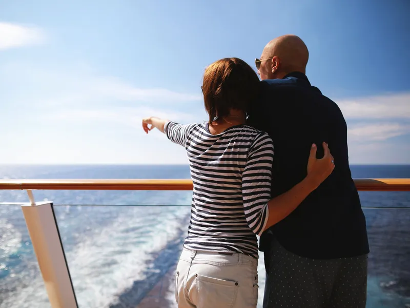 Paar auf Kreuzfahrtschiff blickt auf das Meer und zeigt in die Ferne.