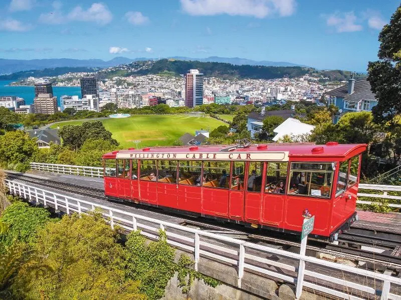 Rote Seilbahn fährt auf Schienen, Skyline und grüne Hügel im Hintergrund.