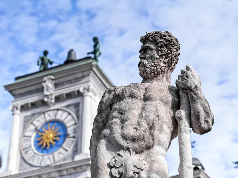 Steinstatue eines bärtigen Mannes vor einem Gebäude mit Uhr und Figuren.