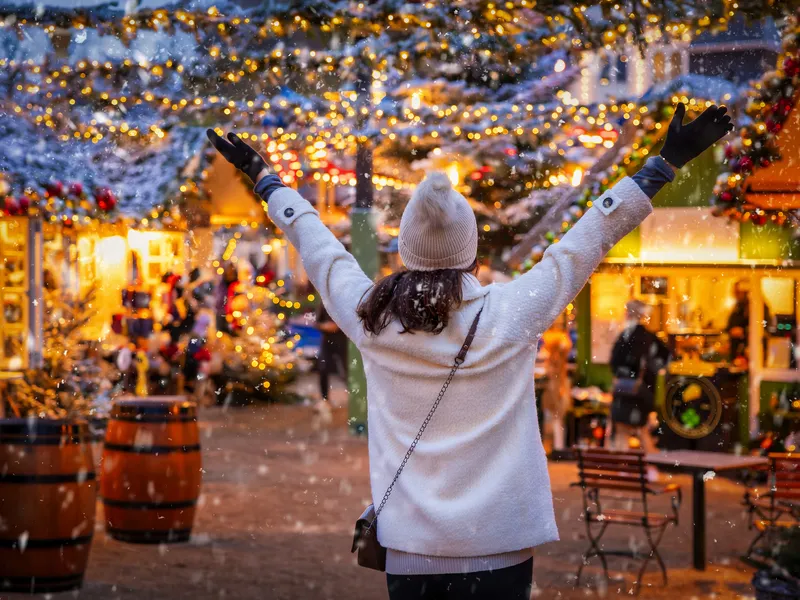 Person auf Weihnachtsmarkt, Hände hoch, umgeben von Lichtern und Dekorationen.