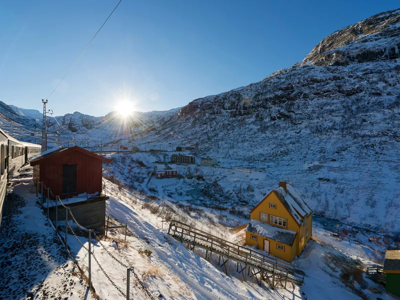 Zug im Schnee mit gelbem Haus und Bergen bei Sonnenschein