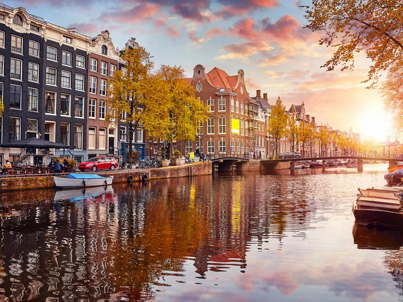 Kanal in Amsterdam bei Sonnenuntergang, mit Booten und herbstlichen Bäumen.