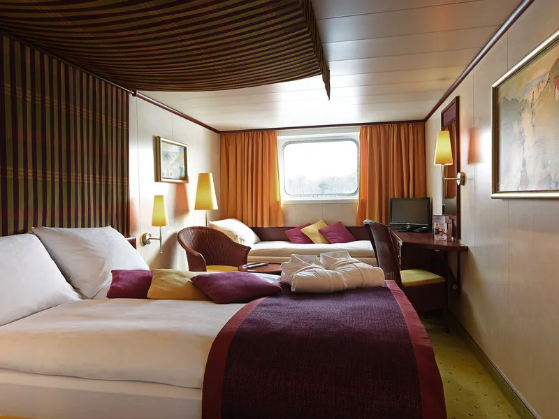 Kabine mit Doppelbett, Zusatzbett, Fenster, Schreibtisch und warmen Farben.