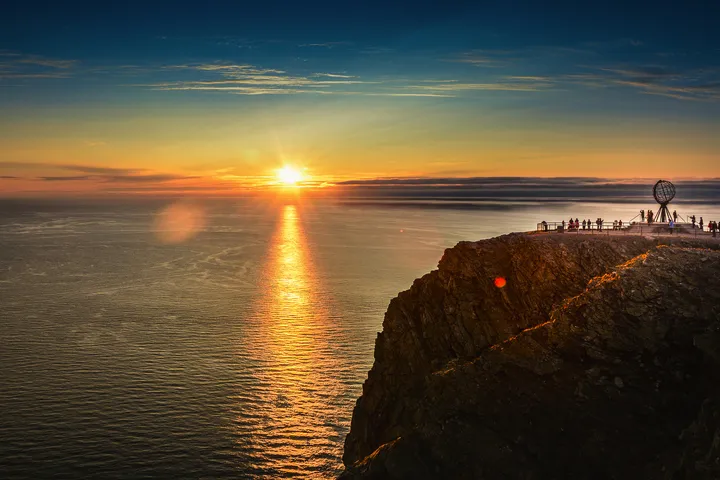 Klippe bei Sonnenuntergang mit Menschen und Globus-Skulptur im Nordkap, Norwegen.