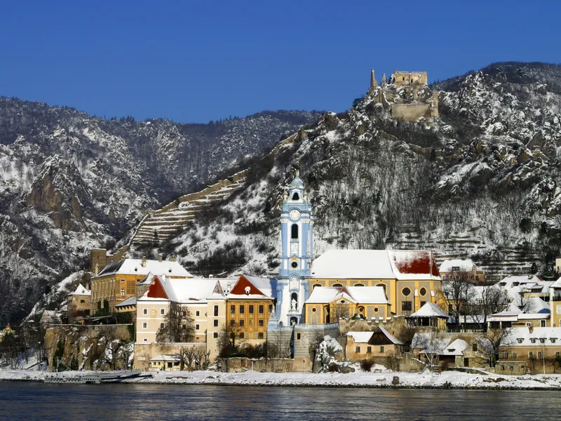 Verschneites Dorf an einem Fluss mit blauer Kirche und Burgruine im Hintergrund.