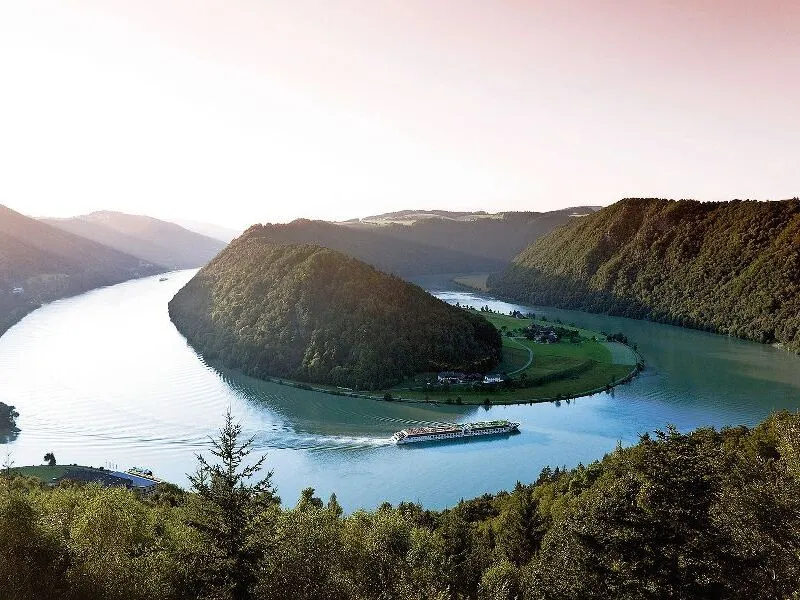 Flusslandschaft mit Schiff in einer Flussschleife, bewaldete Hügel im Hintergrund.